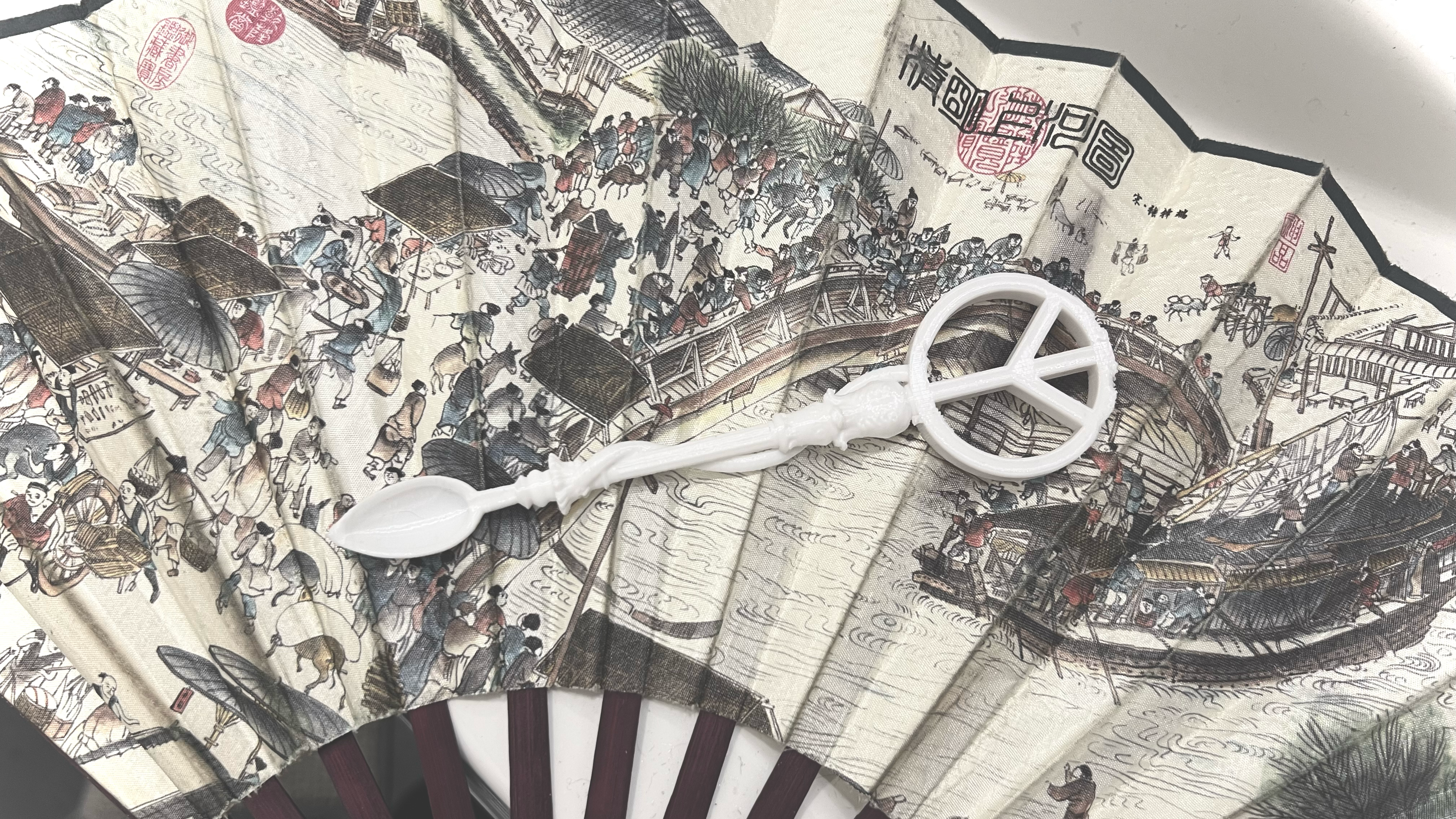 White sculptural spoon with a peace symbol resting on an illustrated folding fan